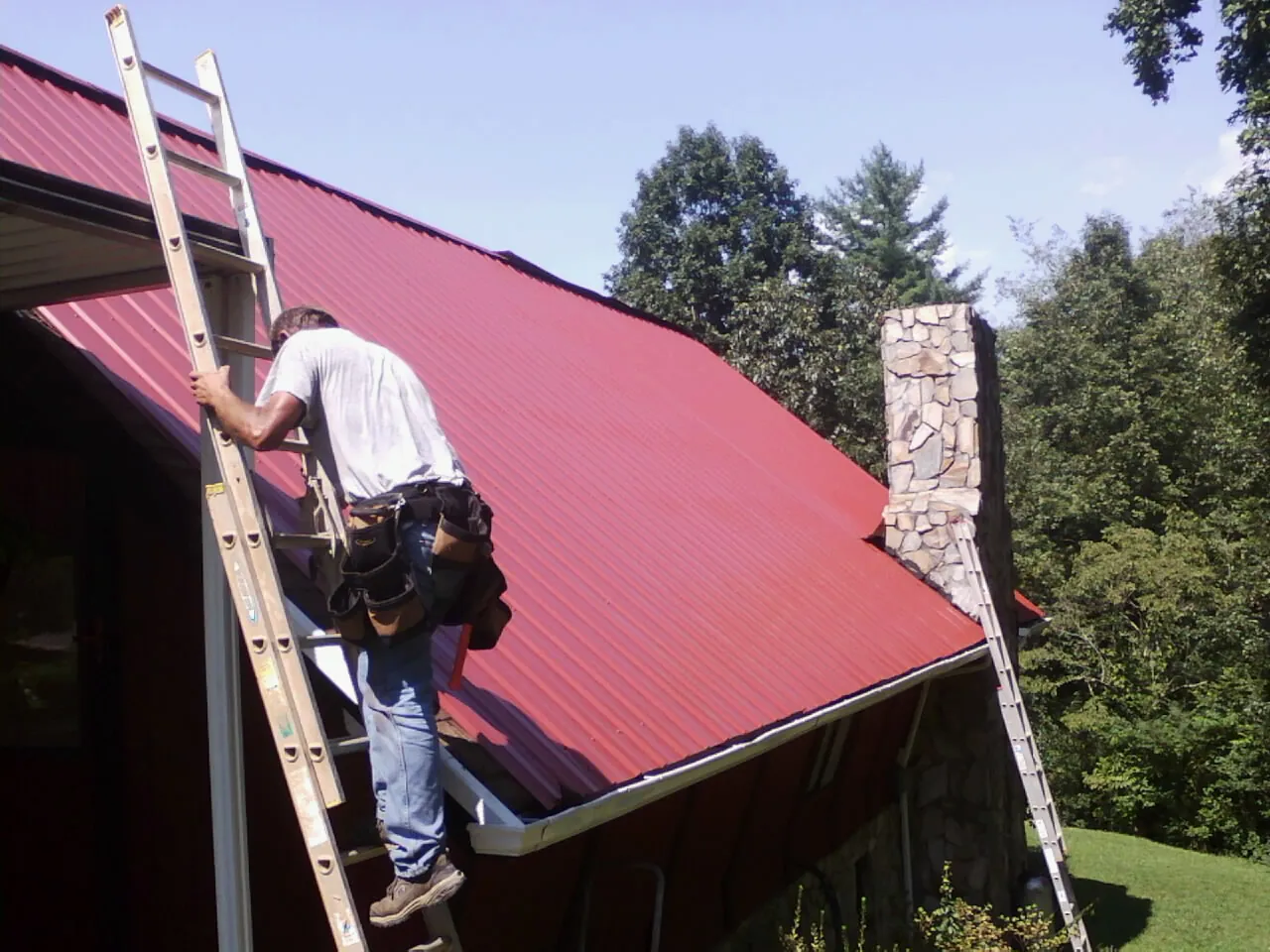 Roofer on a ladder beside a red metal roof and stone chimney — Soffit & Fascia Repair by Roofing Brothers in Beachwood